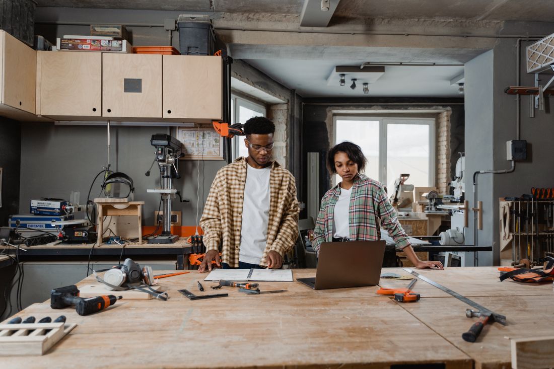 Two people working through something together at a bench, laptop and tools to hand.